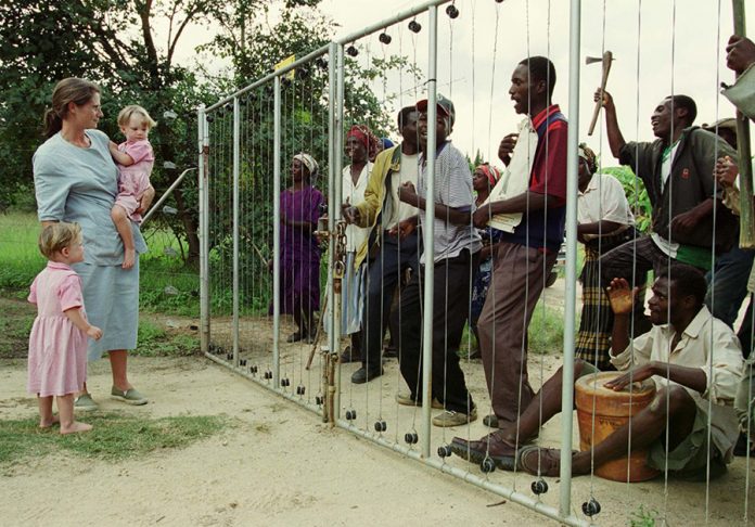 FARMERS WIFE WATCHES FARM INVADERS SING OUTSIDE HER HOMESTEAD IN ZIMBABWE.